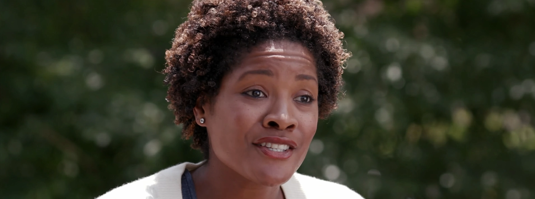 A person with short curly hair speaks outdoors, with a blurred green background.