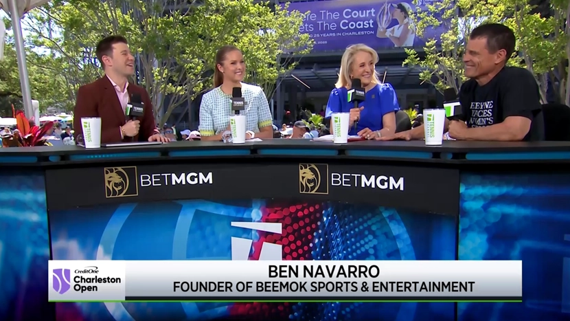 Four people sitting at a broadcast desk outdoors with microphones and branded panels.