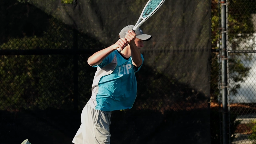 A tennis player in a blue shirt swings a racquet near a court's fence on a sunny day.