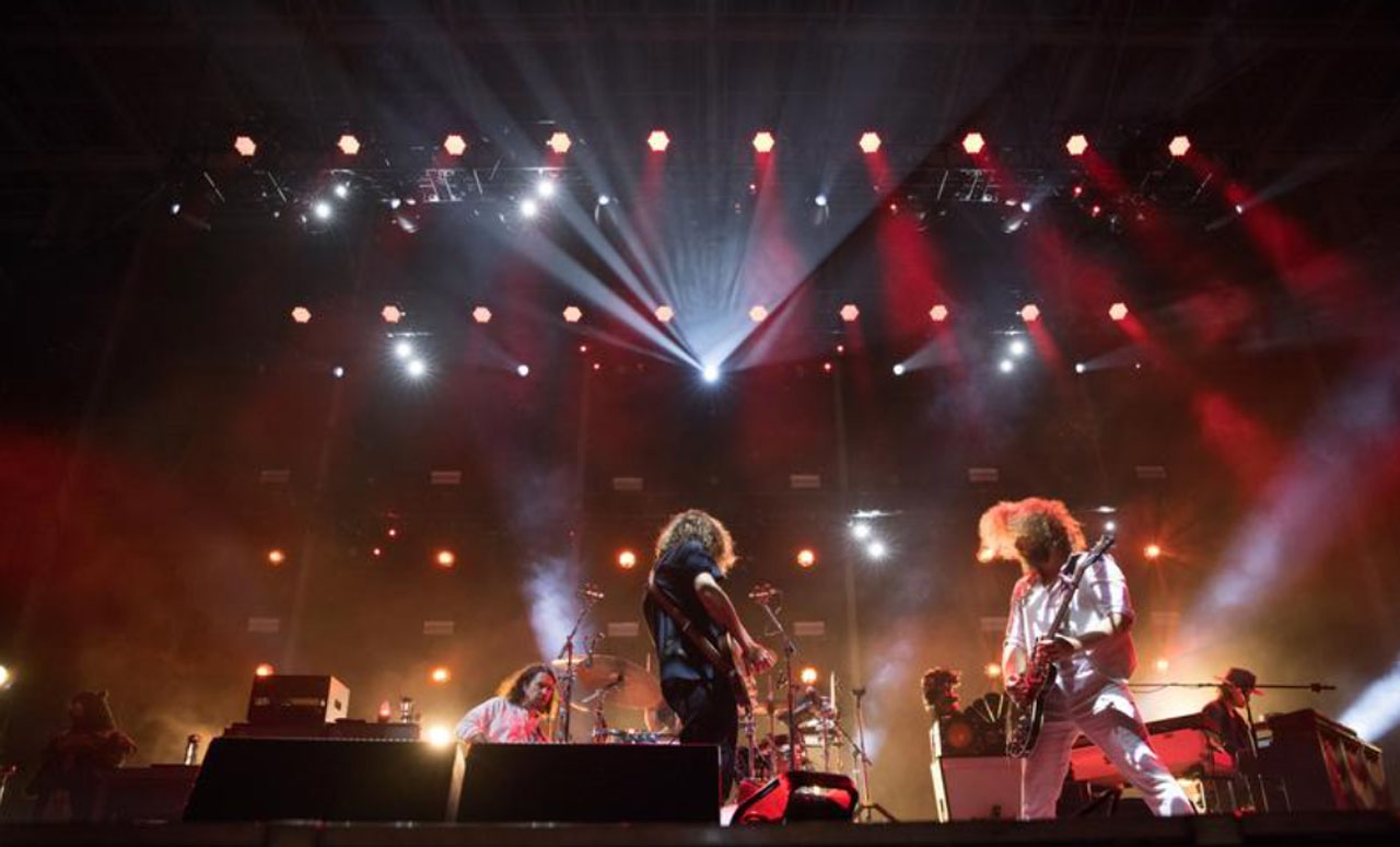 Band performing on stage with vibrant lighting and smoke effects, featuring guitarists and a drummer.