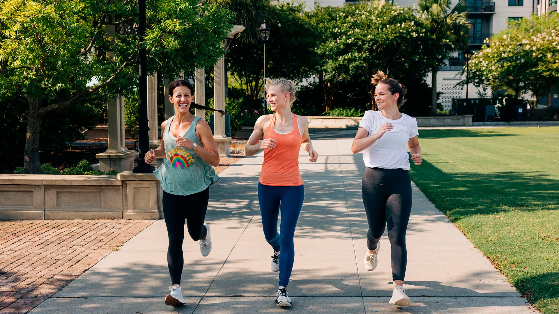 Three people jog along a paved path in a park surrounded by trees and greenery.
