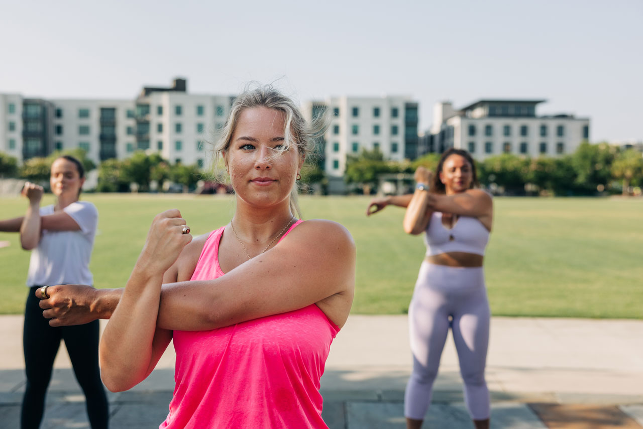Three women are stretching their arms in a park with buildings in the background.