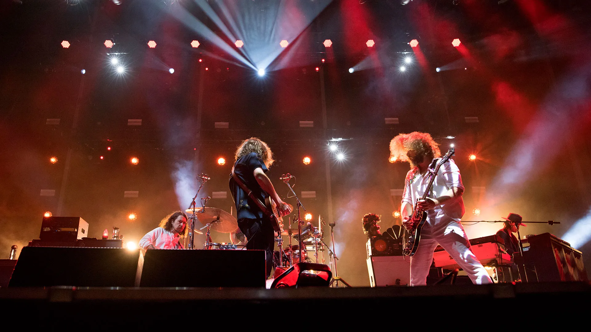 A band performs energetically on stage under bright spotlights, with drums and guitars visible.