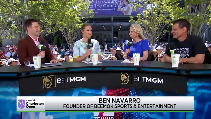 Four people sitting at a broadcast desk outdoors with microphones and branded panels.