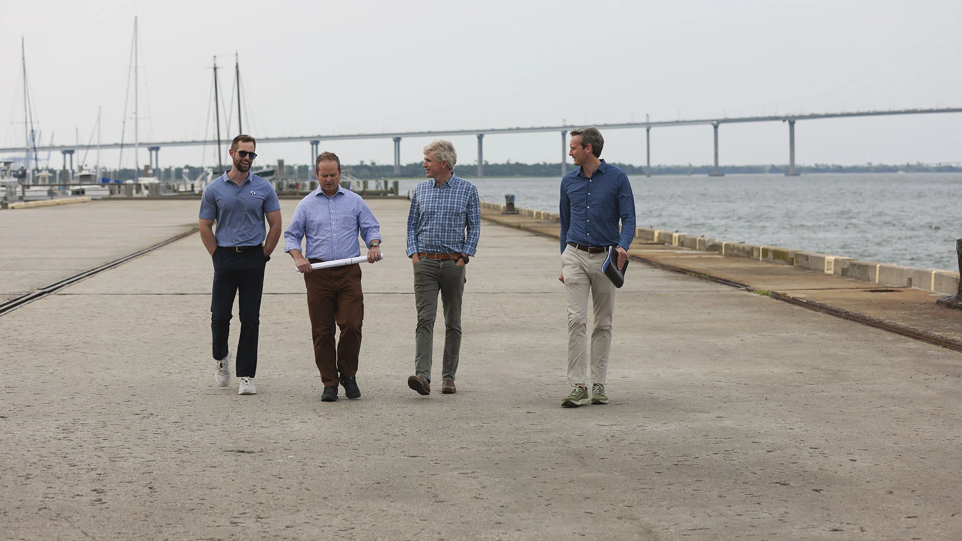 Four men walking along a waterfront dock with a bridge and sailboats in the background.